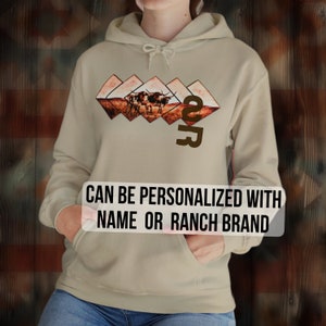 May include: A tan hoodie with a brown graphic of a herd of cattle in a field. The graphic is surrounded by a diamond pattern. The hoodie has a drawstring hood and a kangaroo pocket. The text "2R" is printed on the hoodie. The text "CAN BE PERSONALIZED WITH NAME OR RANCH BRAND" is printed below the graphic.