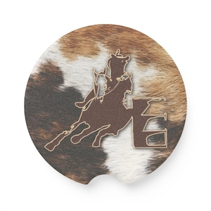 May include: Brown and white cowhide coaster with a silhouette of a barrel racer on a horse jumping over a barrel. The barrel is labeled with the letter "E".