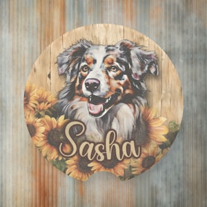 May include: A round wooden coaster with a painted image of an Australian Shepherd dog with brown, white, and black fur. The dog is surrounded by sunflowers and the name "Sasha" is written in a cursive font.
