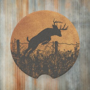 May include: A round car coaster with a deer jumping over a fence silhouette design. The coaster has a textured surface with a brown and black color scheme. The background is a gradient of orange and brown.
