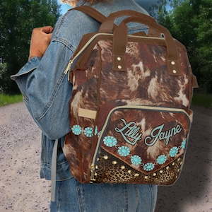 May include: Brown and white cowhide backpack with a brown leather handle and a zipper closure. The backpack has a large pocket on the front with a brown and black leopard print fabric and gold studs. The pocket has the text "Lilly Jayne" embroidered in white with turquoise blue embellishments.
