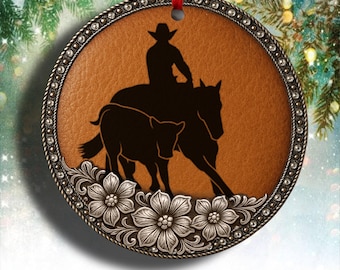 Pode incluir: Enfeite de Natal redondo com fundo castanho semelhante a couro. Apresenta uma silhueta preta de um cowboy a cavalo, pastoreando um bezerro. O enfeite tem uma borda floral prateada decorativa e uma fita vermelha para pendurar.