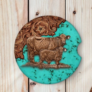 May include: A turquoise and brown coaster with a design of a cow and calf. The cow and calf are standing on a green field with a brown leather background.