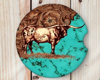 May include: Round car coaster featuring a longhorn steer illustration. The coaster has a turquoise background with a brown leather-like floral design and a brown steer illustration. The coaster is designed to fit in a car's cup holder.