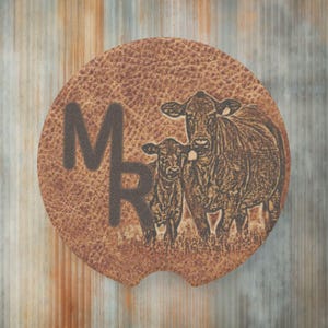 May include: Brown leather coaster with a design of two cows, one larger than the other, with the letters "MR" in the upper left corner.