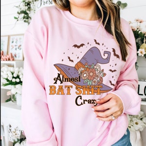 May include: A light pink sweatshirt featuring a witch's hat with floral accents and bats. The text on the sweatshirt reads "Almost BAT SHIT Crazy" in a retro font. The design is centered on the front of the sweatshirt.
