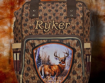 May include: A brown and tan backpack with a deer print design. The backpack has a large front pocket with a deer image and the name "Ryker" embroidered on the top flap.