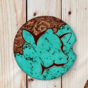 May include: Round coaster with a turquoise blue background and a brown leather design. The design features a silhouette of a cow's head.