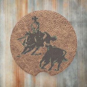 May include: A brown leather coaster with a silhouette of a cowboy riding a bucking bronco. The cowboy is wearing a hat and the bronco is rearing up.
