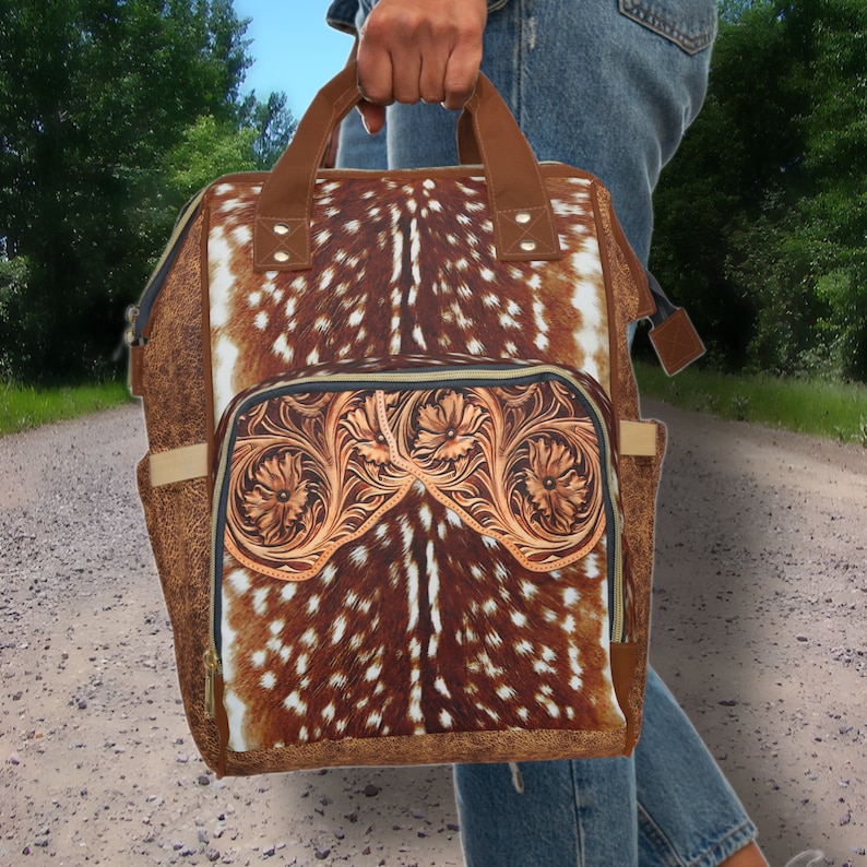 May include: A brown leather backpack with a deer print design and tooled leather accents. The backpack has a large main compartment and a smaller front pocket.