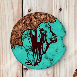 May include: A round turquoise and brown coaster with a silhouette of a steer in the center. The coaster has a decorative floral pattern around the edge.