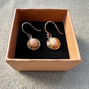 May include: A pair of silver earrings featuring a small, oval-shaped seashell set in a gold-toned surround. The earrings are presented in a brown box with a black lining.