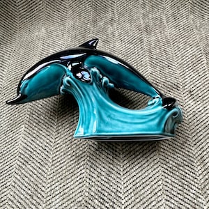 May include: A ceramic dolphin figurine in teal and black. The dolphin is arched, leaping over a teal wave. The figurine has a glossy finish and is a decorative item.