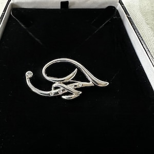 Puede incluir: Un broche de plata en forma de letra A estilizada, con pequeños diamantes de imitación transparentes engastados en el diseño.