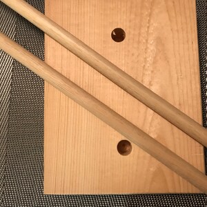 May include: Two wooden dowels laying on top of a light wood board with two holes drilled in it.