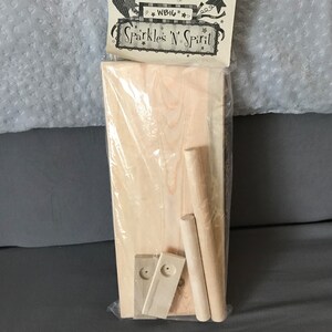 May include: A packaged wooden craft kit with the text "Sparkles 'N' Spirit". The kit includes a rectangular wooden board, two cylindrical dowels, and two smaller wooden pieces, all in natural wood tones. The kit is packaged in clear plastic.