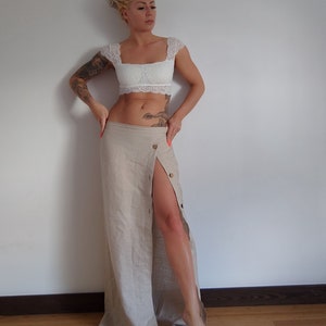 May include: A woman wearing a white lace crop top and a beige linen skirt with a side slit and a button closure. She has blonde hair and tattoos on her arms.