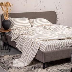 May include: A grey linen bed with white stripes. The bed is made with a white and grey striped duvet cover and two matching pillowcases. The bed is sitting on a grey rug with a black and white pattern.