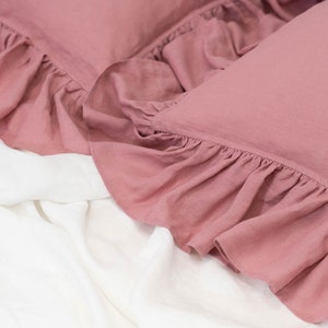 Puede incluir: Dos fundas de almohada de color rosa polvoriento con bordes con volantes, sobre un cubrecama blanco.