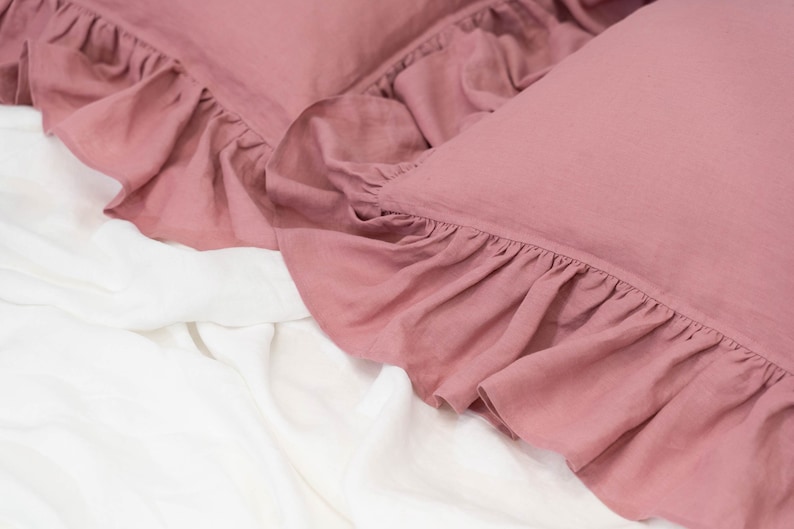 Puede incluir: Dos fundas de almohada de color rosa polvoriento con bordes con volantes, sobre un cubrecama blanco.