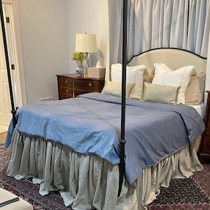 May include: A blue and beige bed skirt with a ruffled edge. The bed skirt is on a bed with a blue duvet cover and white and beige pillows. The bed is in a bedroom with a wooden dresser and a lamp.