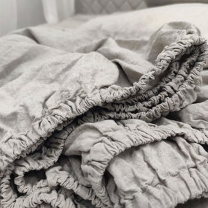 Peut inclure: Un drap-housse gris clair avec des bords élastiques, fabriqué dans un tissu doux et texturé. Le drap est légèrement froissé, suggérant du lin naturel ou une matière similaire. L'arrière-plan comprend un oreiller et d'autres éléments de literie.