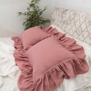 Puede incluir: Dos fundas de almohada de lino rosa polvoriento con bordes con volantes. Las fundas de almohada est&aacute;n sobre una cama blanca con una cabecera acolchada.