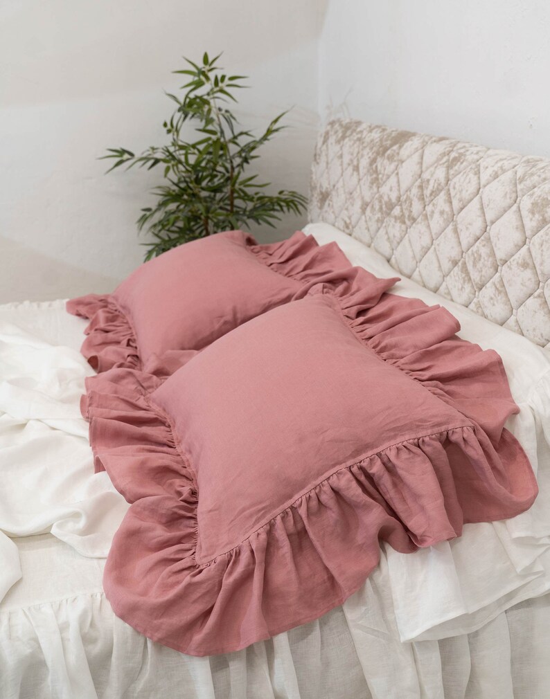 Puede incluir: Dos fundas de almohada de lino rosa polvoriento con bordes con volantes. Las fundas de almohada est&aacute;n sobre una cama blanca con una cabecera acolchada.