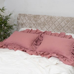 Puede incluir: Dos fundas de almohada de color rosa polvoriento con volantes sobre una cama blanca con una cabecera capiton&eacute; beige.
