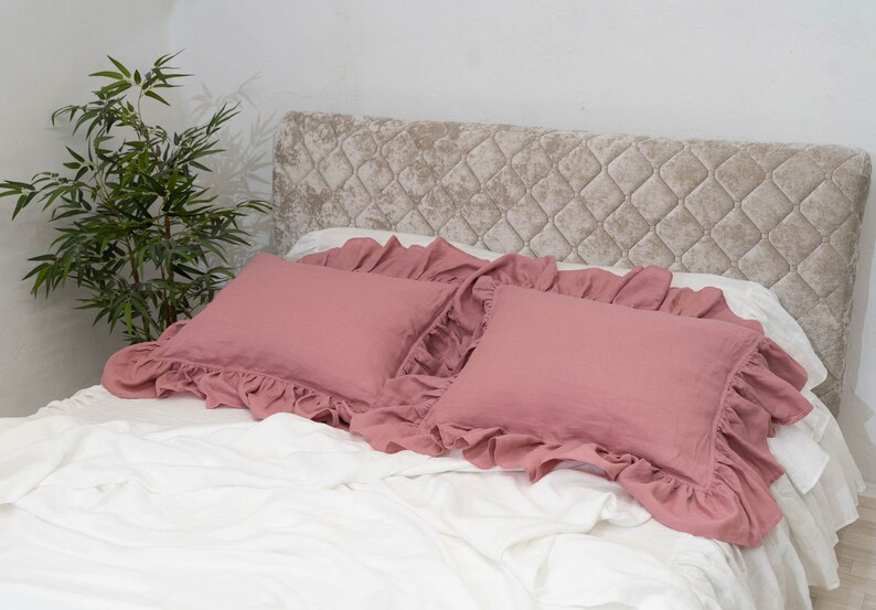 Puede incluir: Dos fundas de almohada de color rosa polvoriento con volantes sobre una cama blanca con una cabecera capiton&eacute; beige.