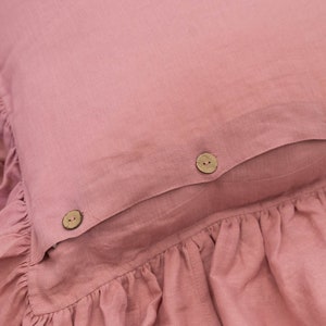 Puede incluir: Una almohada rosa polvorienta con un borde con volantes y tres botones de madera. La almohada est&aacute; hecha de una tela suave de lino.