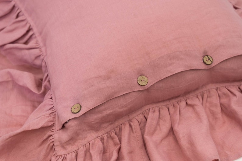 Puede incluir: Una almohada rosa polvorienta con un borde con volantes y tres botones de madera. La almohada est&aacute; hecha de una tela suave de lino.