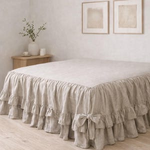May include: A beige linen bed skirt with a ruffled design and bow accents. The bed skirt covers the base of the bed, adding a decorative element to the bedroom. The neutral color complements various bedroom styles.