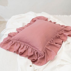 Puede incluir: Una almohada rosa polvorienta con un borde con volantes. La almohada est&aacute; hecha de una tela de lino suave y est&aacute; sentada sobre una colcha blanca.