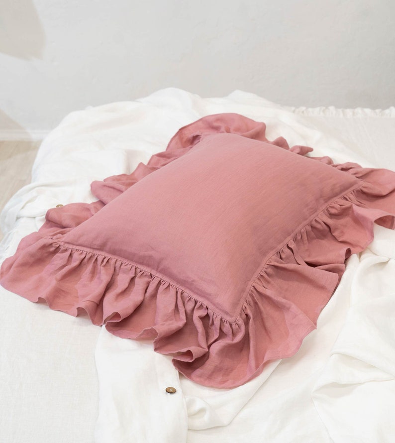 Puede incluir: Una almohada rosa polvorienta con un borde con volantes. La almohada est&aacute; hecha de una tela de lino suave y est&aacute; sentada sobre una colcha blanca.