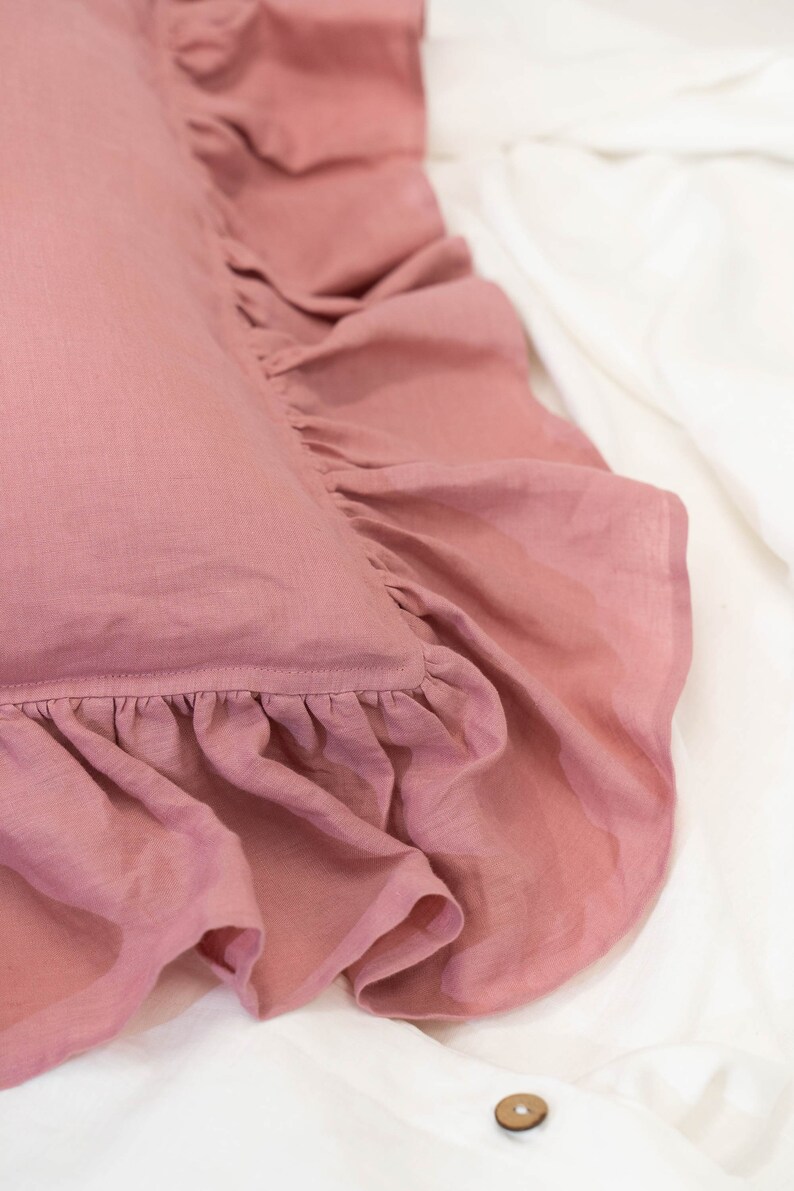 Puede incluir: Una almohada rosa polvorienta con un borde con volantes. La almohada est&aacute; hecha de una tela de lino suave.