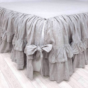 May include: A gray linen bed skirt with a ruffled edge and a bow at the center. The skirt has multiple layers of ruffles.