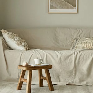 Puede incluir: Un salón de tonos neutros con un sofá beige cubierto con una funda a juego. Cojines decorativos con detalles texturizados descansan sobre el sofá. Una pequeña mesa de madera sostiene una taza y un libro. El ambiente es minimalista y acogedor.