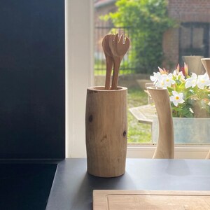 Peut inclure: Un porte-ustensiles en bois avec une finition en bois naturel. Le porte-ustensiles est rempli d'ustensiles en bois, y compris des fourchettes et des cuillères. Le porte-ustensiles est posé sur un comptoir noir devant une fenêtre.