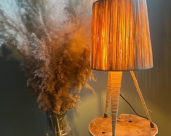 May include: A woven lampshade with a light brown and white color scheme sits on a wooden tripod stand. The lamp is on and casting a warm glow. The lamp is next to a tall glass vase filled with dried pampas grass.