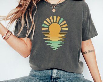 Puede incluir: Camiseta gris oscuro con un gráfico de puesta de sol. El diseño incluye un sol amarillo con rayos naranjas y verdes, reflejado en el agua. La camiseta es de corte holgado y tiene mangas cortas.