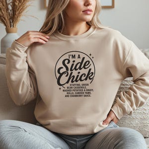 May include: Beige crew neck sweatshirt featuring the text "I'M A Side Chick" in a black circular design. The design lists Thanksgiving side dishes. A casual, comfortable top for the holiday season.
