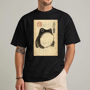May include: Black t-shirt featuring a vintage-style illustration of a frog with a large open mouth. The frog is in black, with a beige background and Japanese text. The shirt is a classic crew neck.
