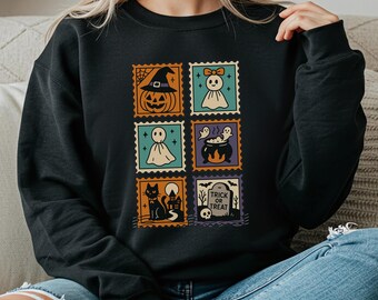 Può includere: Felpa nera con sei disegni di francobolli a tema Halloween. I francobolli raffigurano una zucca, un fantasma, un calderone, un gatto nero e una lapide con la scritta "Trick or Treat". I disegni sono in arancione, verde acqua e viola.
