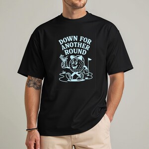 May include: Black t-shirt featuring a cartoon golf ball character with a golf club, cocktail, and the text "DOWN FOR ANOTHER ROUND" in light blue. The golf ball is wearing a golf hat and sitting on grass.