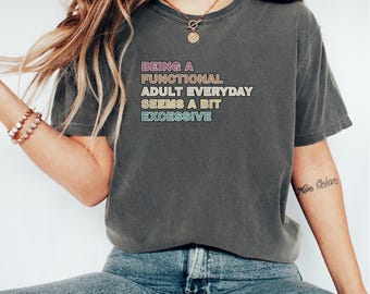 May include: Dark gray t-shirt with the text "BEING A FUNCTIONAL ADULT EVERYDAY SEEMS A BIT EXCESSIVE" in pastel colors. The shirt has a crew neck and short sleeves. The model is wearing blue jeans and a gold necklace.