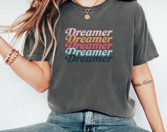 May include: Dark grey t-shirt featuring the word "Dreamer" stacked five times in a retro font. The text colours are pink, orange, red, blue, and teal. The shirt is worn with blue jeans.