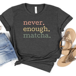 以下が含まれることがあります： 「never. enough. matcha.」とピンク、黄色、緑色の文字がプリントされた濃いグレーのTシャツ。デニムショートパンツとゴールドのサンダルと合わせています。