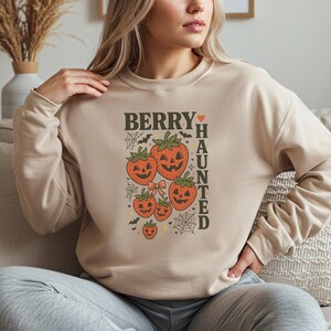 May include: Beige sweatshirt with a Halloween-themed graphic. The design features smiling jack-o'-lantern strawberries, spiderwebs, and the text "BERRY HAUNTED". A fun and festive top for the spooky season.