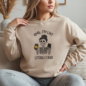 Puede incluir: Sudadera beige con un gráfico negro de un esqueleto sosteniendo una taza de café y un teléfono. El texto sobre el esqueleto dice "OMG, I'M LIKE" y debajo "LITERALLY DEAD".